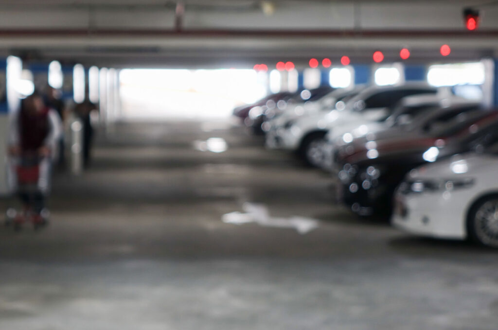 Blurred view of cars parked in an indoor parking garage with red overhead lights and pedestrians walking between vehicles.
