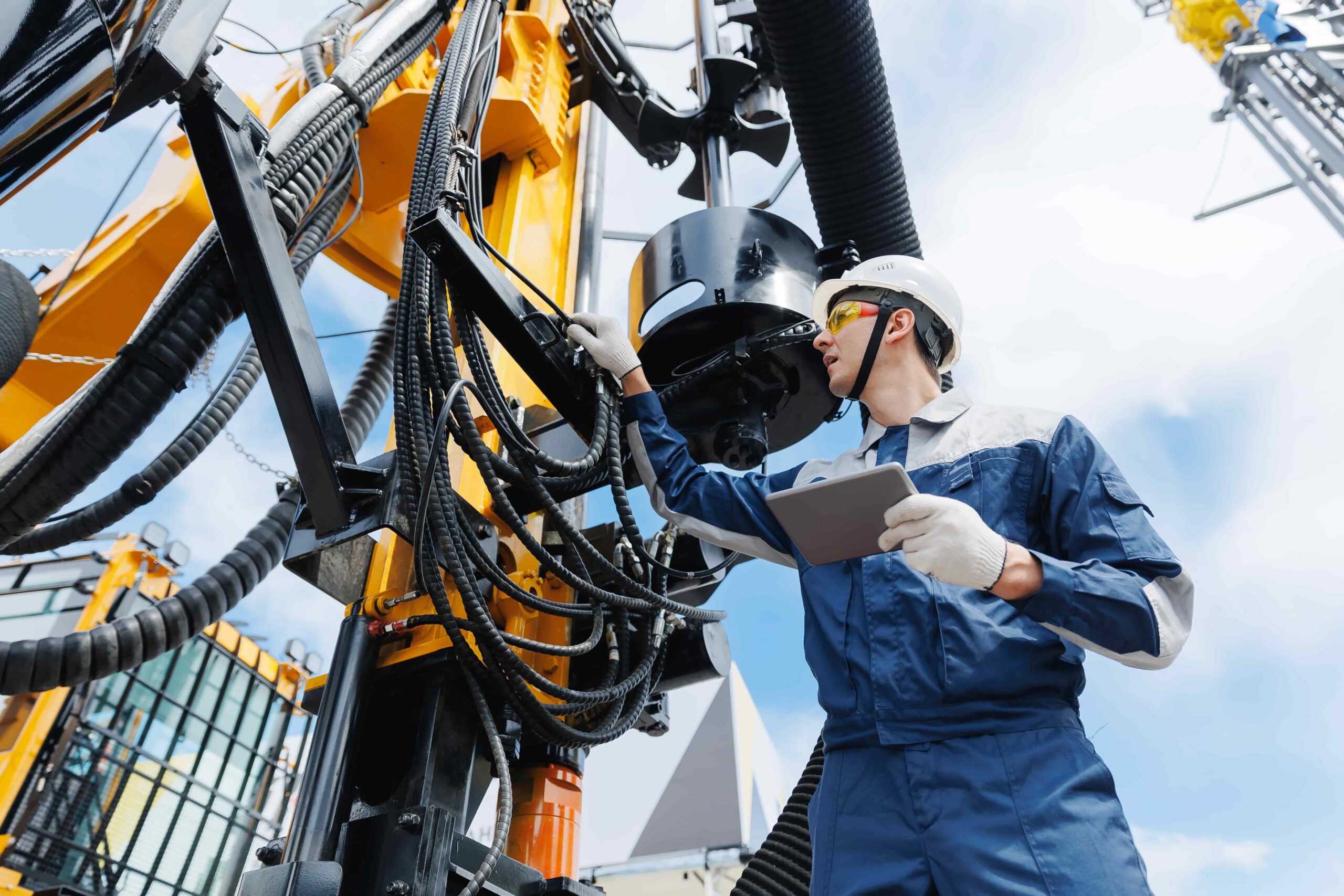 Industrial worker in safety gear inspecting heavy drilling machinery with tablet, representing equipment maintenance and workplace safety in construction or oil industry