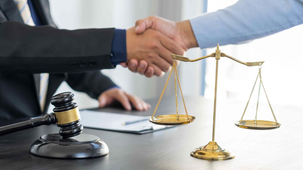 Two professionals shaking hands over a desk with a judge&rsquo;s gavel and scales of justice, symbolizing a legal agreement, settlement, or successful law consultation.