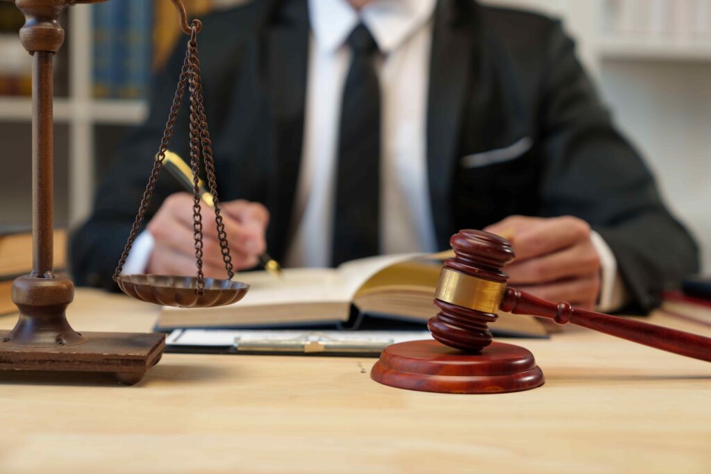 Judge’s gavel and scales of justice on a desk with a lawyer writing in a law book, symbolizing legal proceedings, justice, and courtroom decision-making. Judge’s gavel and scales of justice on a desk with a lawyer writing in a law book, symbolizing legal proceedings, justice, and courtroom decision-making.