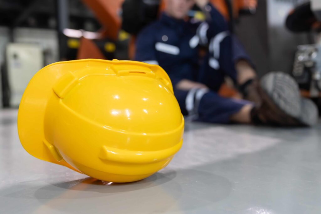 Yellow safety hard hat in foreground with injured worker lying on factory floor, highlighting workplace accident and occupational safety risk